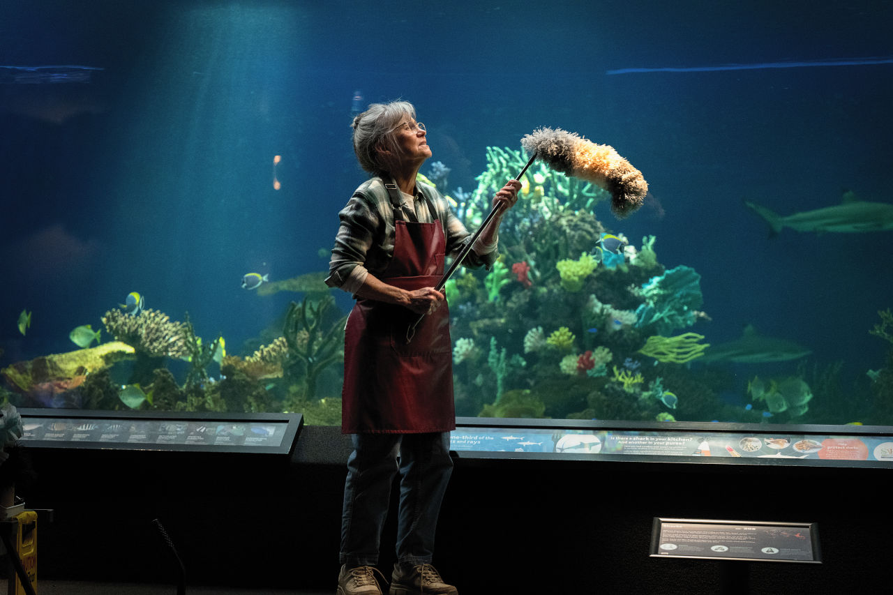 Tova sitting beside a giant Pacific octopus exhibit inside an aquarium, surrounded by glowing underwater displays